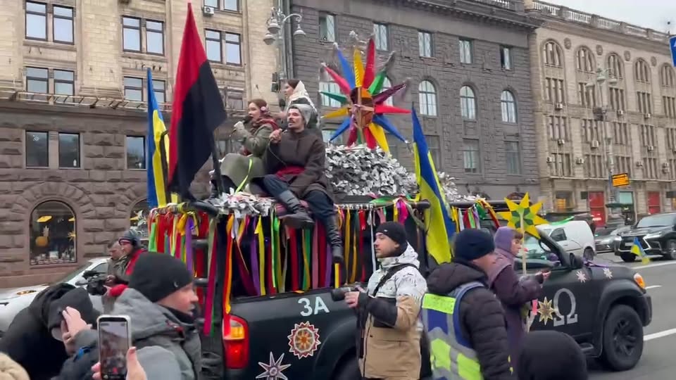 People sing from a pickup decorated with ribbons, Ukrainian flags and colourful star ornaments, surrounded by people in winter coats.