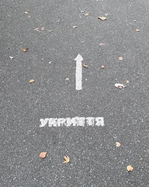 A white arrow and the Ukrainian word for shelter painted on grey pavement scattered with fallen leaves.